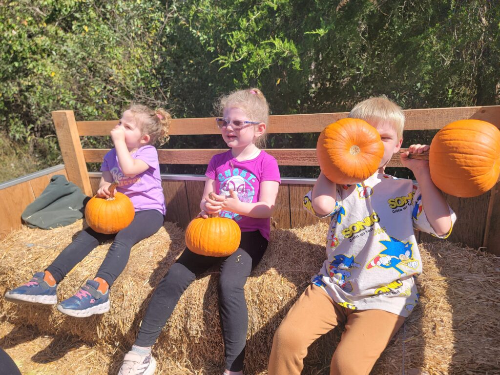 Three children sitting on hay bales inside the Timber Valley Trailblazer wagon, each holding a pumpkin after a fun day of picking at Timber Valley Retreat.