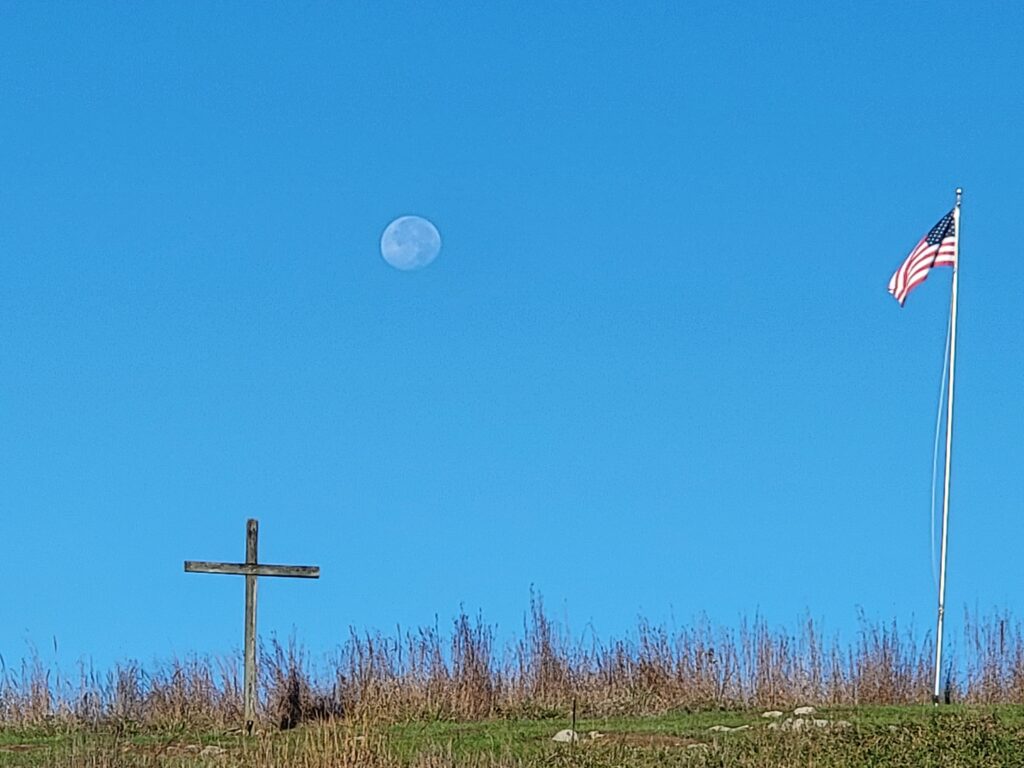 Daytime moon above a hillside with a cross and American flag at Timber Valley Retreat